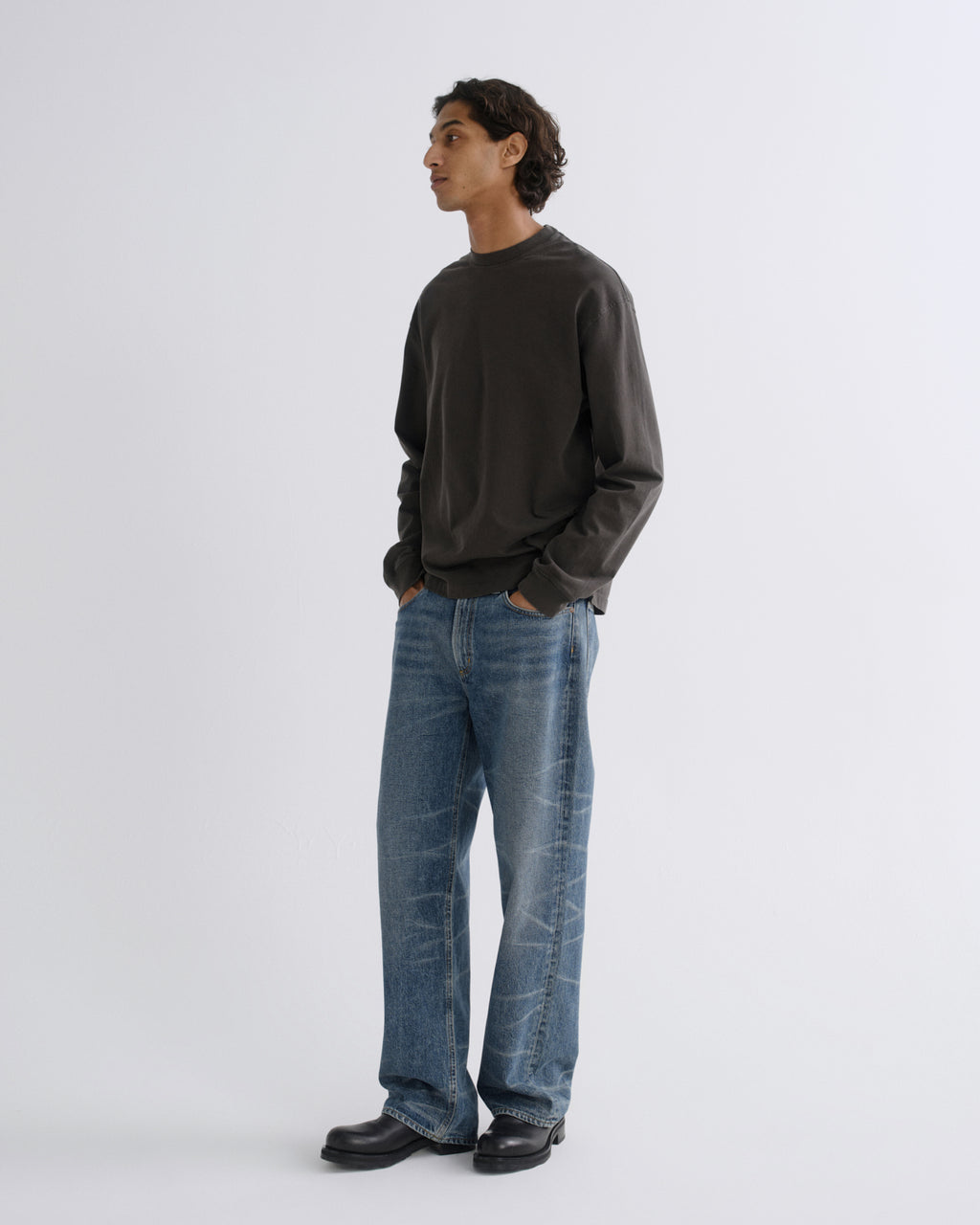 A man wearing a dark charcoal crewneck sweatshirt, blue wide-leg jeans, and black leather shoes, standing in profile against a light gray studio background.