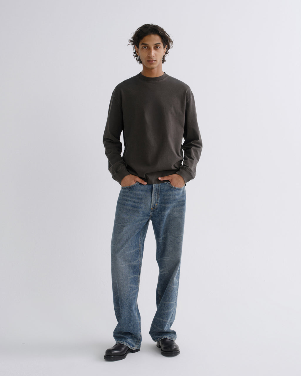 A man stands against a light gray studio background wearing a dark charcoal long-sleeve crewneck sweatshirt, blue jeans, and black shoes.