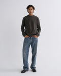 A man stands against a light gray studio background wearing a dark charcoal long-sleeve crewneck sweatshirt, blue jeans, and black shoes.