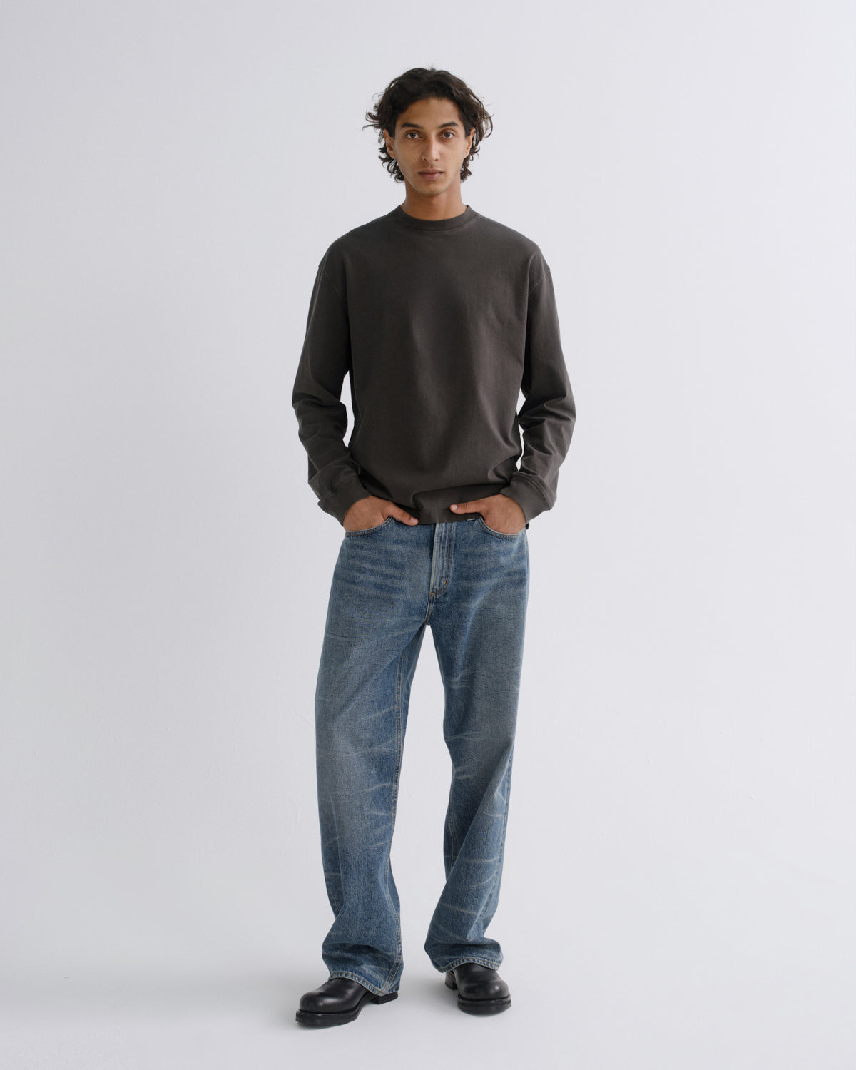 A man stands against a light gray studio background wearing a dark charcoal long-sleeve crewneck sweatshirt, blue jeans, and black shoes.