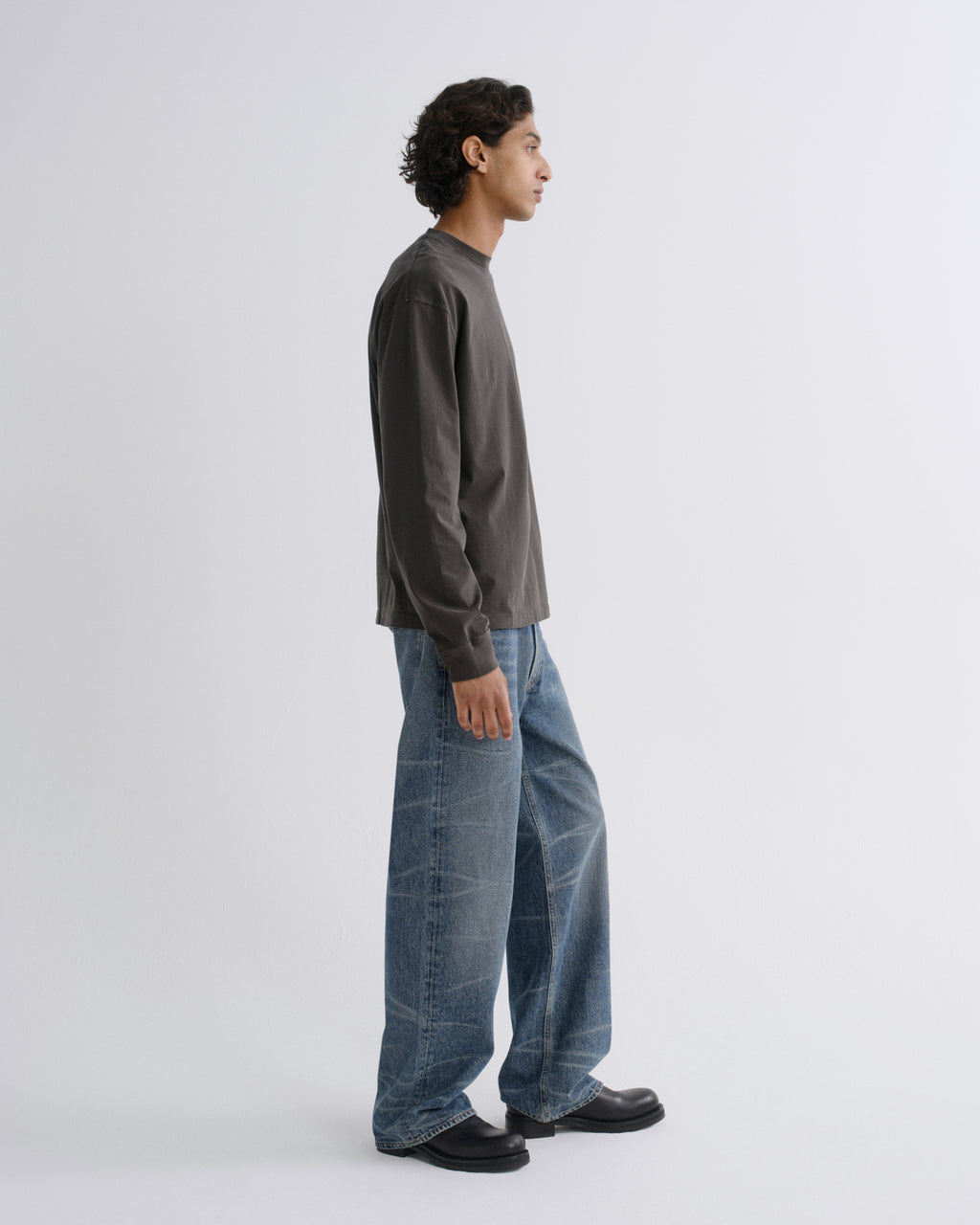 A man in profile wearing a dark charcoal gray long-sleeve shirt, wide-leg blue jeans, and black loafers, standing against a light gray studio background.