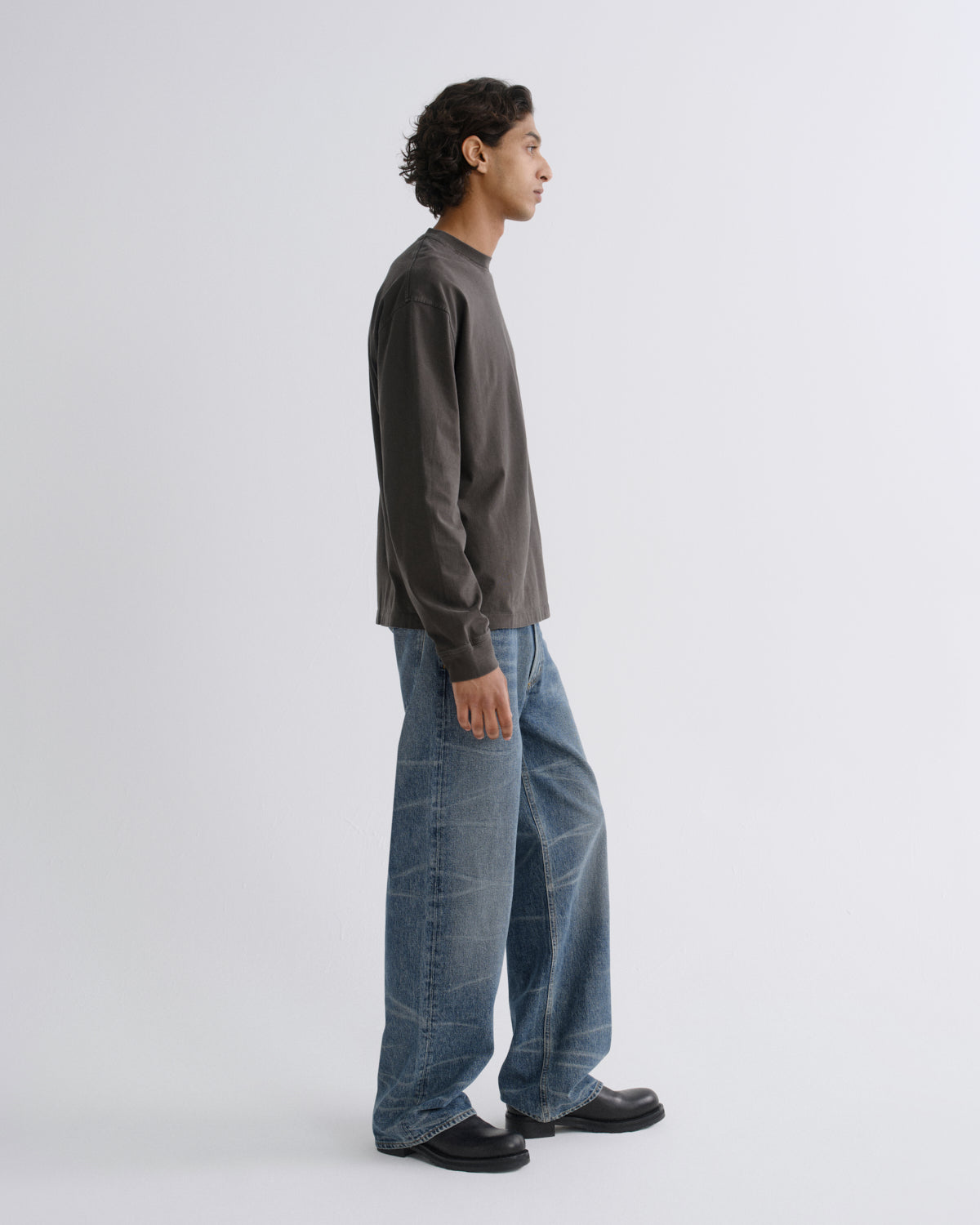 A man in profile wearing a dark charcoal gray long-sleeve shirt, wide-leg blue jeans, and black loafers, standing against a light gray studio background.