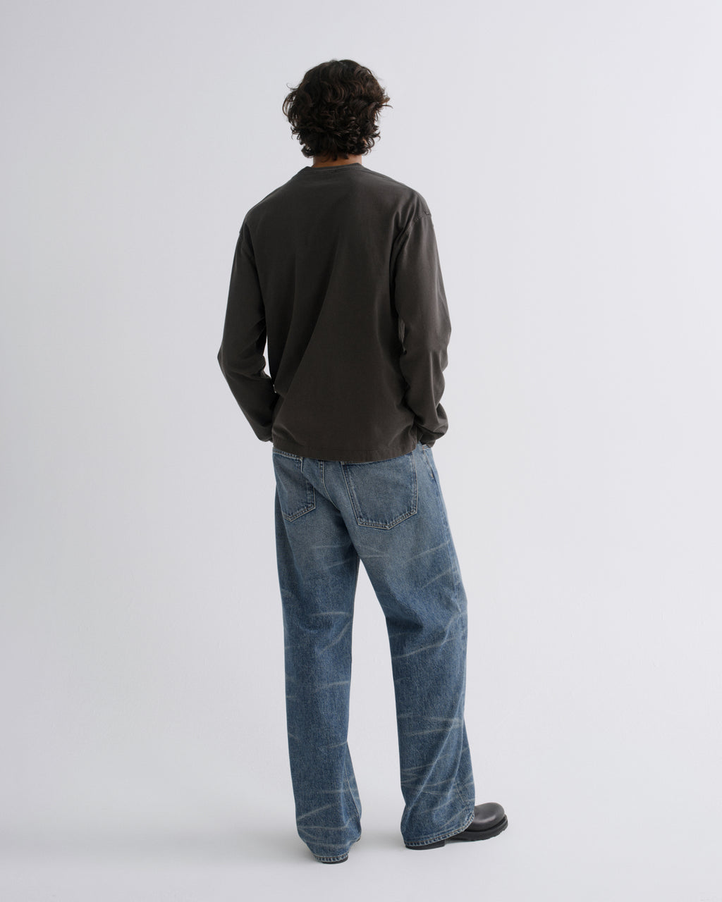 A man viewed from behind wearing a dark charcoal long-sleeve shirt and blue jeans, standing against a light gray studio background.
