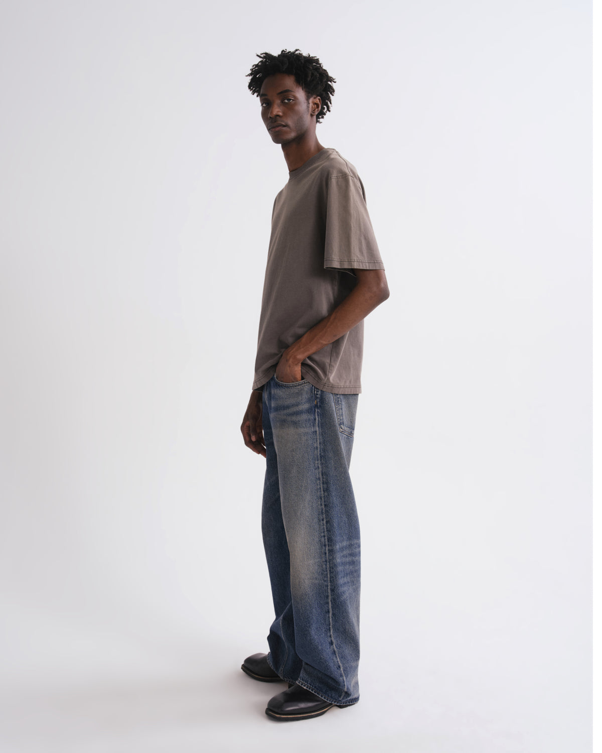 A man wearing a brown oversized t-shirt and wide-leg blue jeans, standing in profile against a plain white studio background.