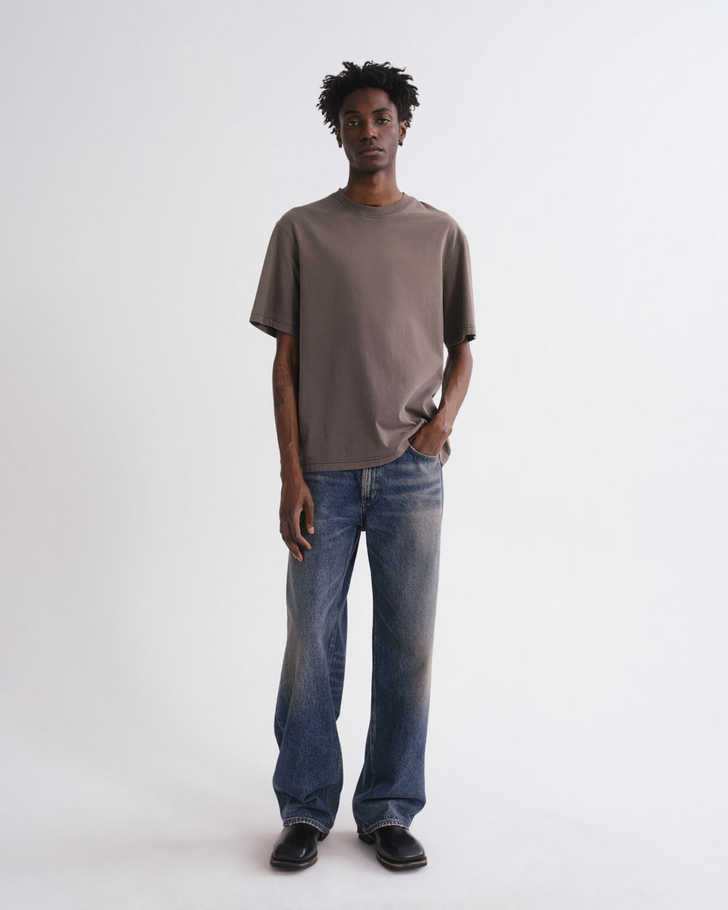 A man modeling an oversized taupe-brown short-sleeve t-shirt and wide-leg blue jeans against a plain white studio background.