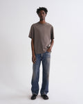A man modeling an oversized taupe-brown short-sleeve t-shirt and wide-leg blue jeans against a plain white studio background.