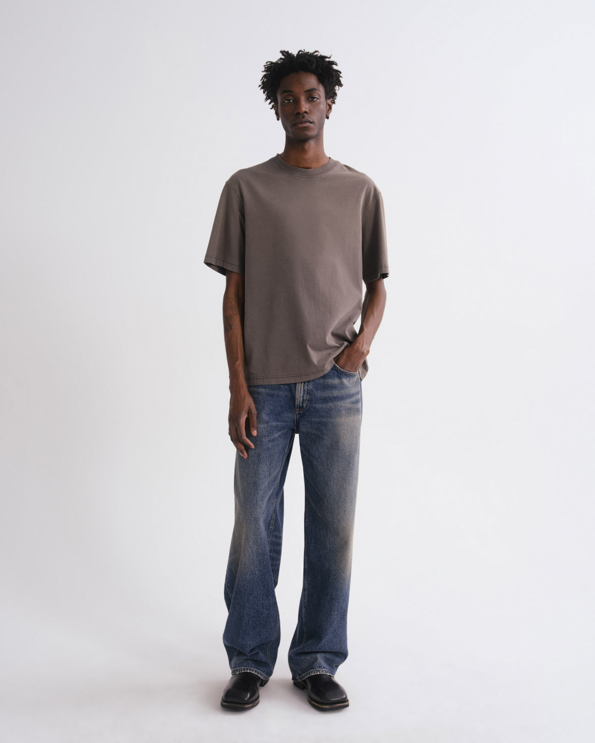 A man modeling an oversized taupe-brown short-sleeve t-shirt and wide-leg blue jeans against a plain white studio background.