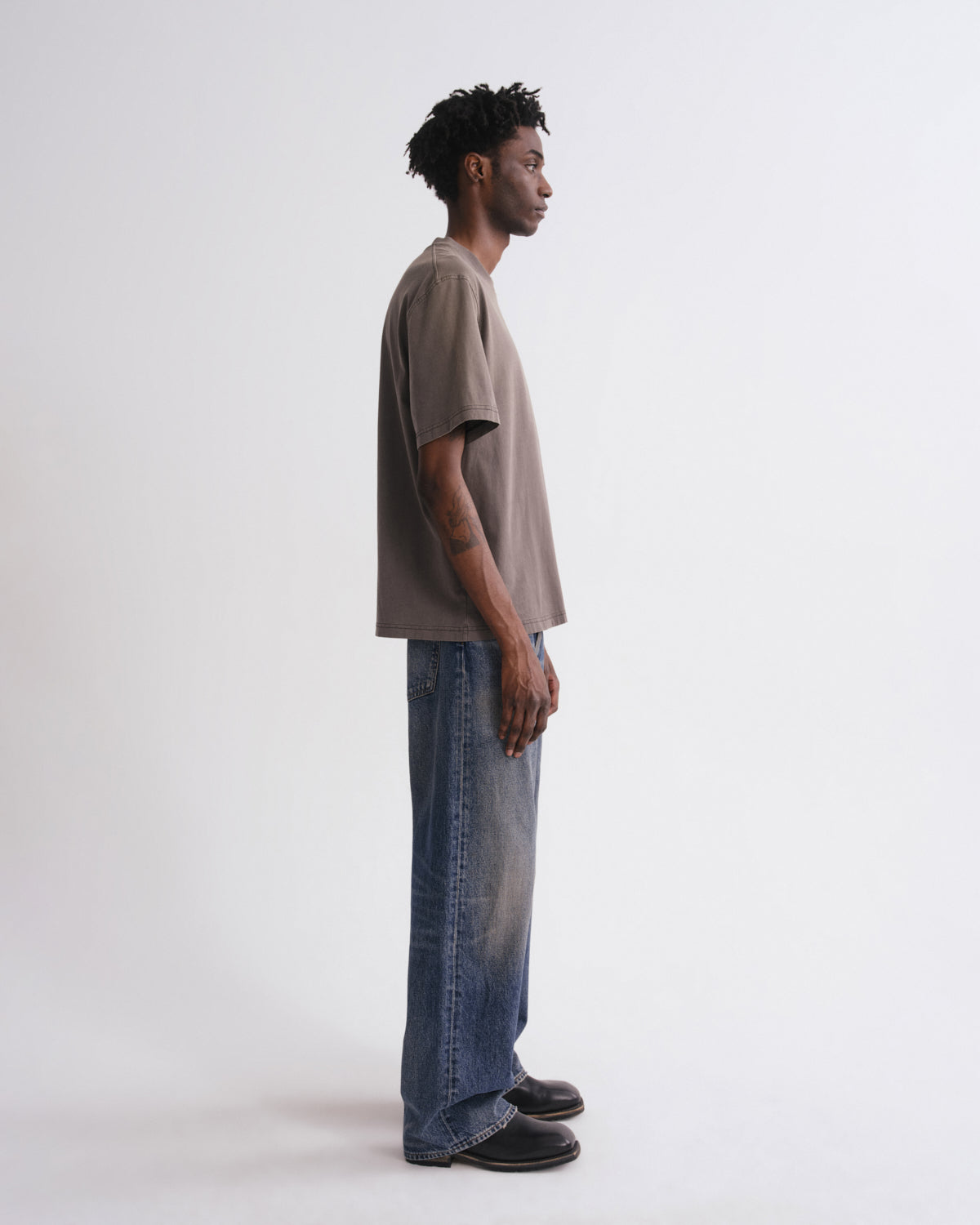A man in profile wearing a taupe oversized short-sleeve t-shirt, wide-leg faded blue jeans, and black shoes, standing against a plain white studio background.