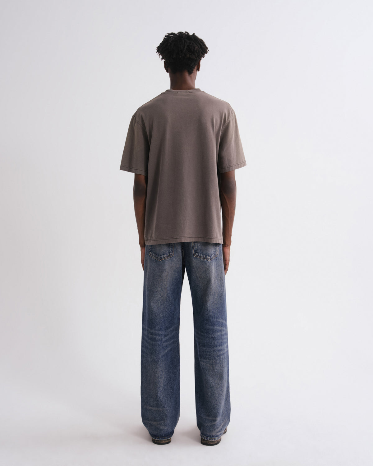 A man viewed from behind wearing an oversized taupe-brown t-shirt and wide-leg blue jeans, standing against a plain white studio background.