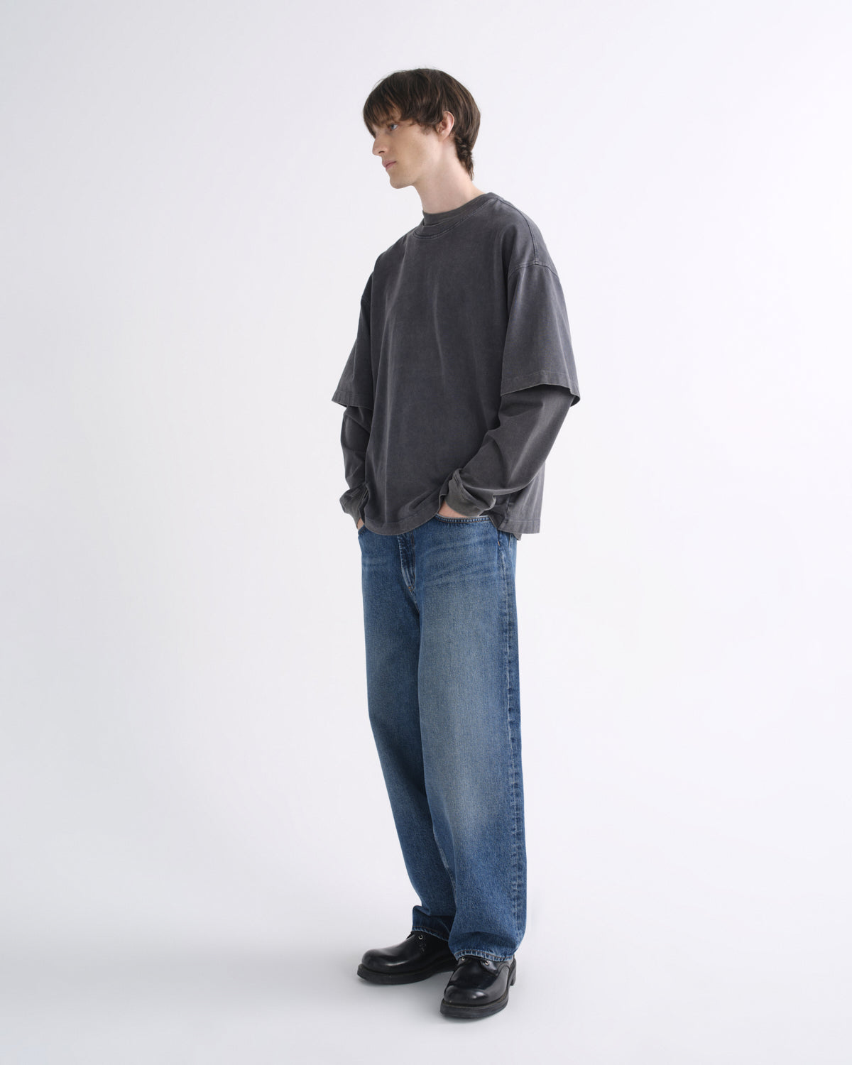 Person wearing a dark grey short sleeve T-shirt layered over a black long sleeve shirt with blue jeans and black shoes on a grey background