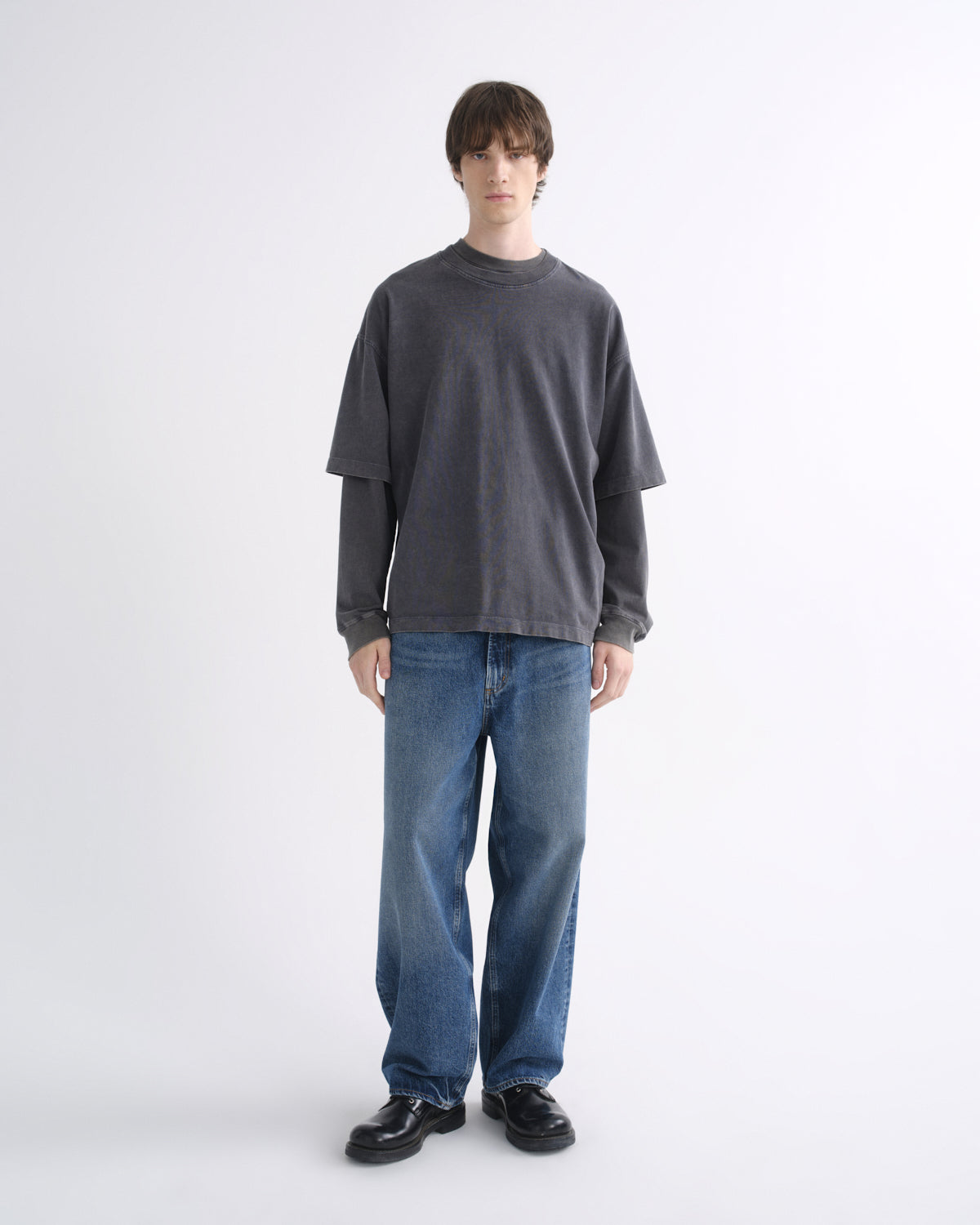 Person wearing a dark grey short sleeve T-shirt layered over a black long sleeve shirt with blue jeans and black shoes on a grey background