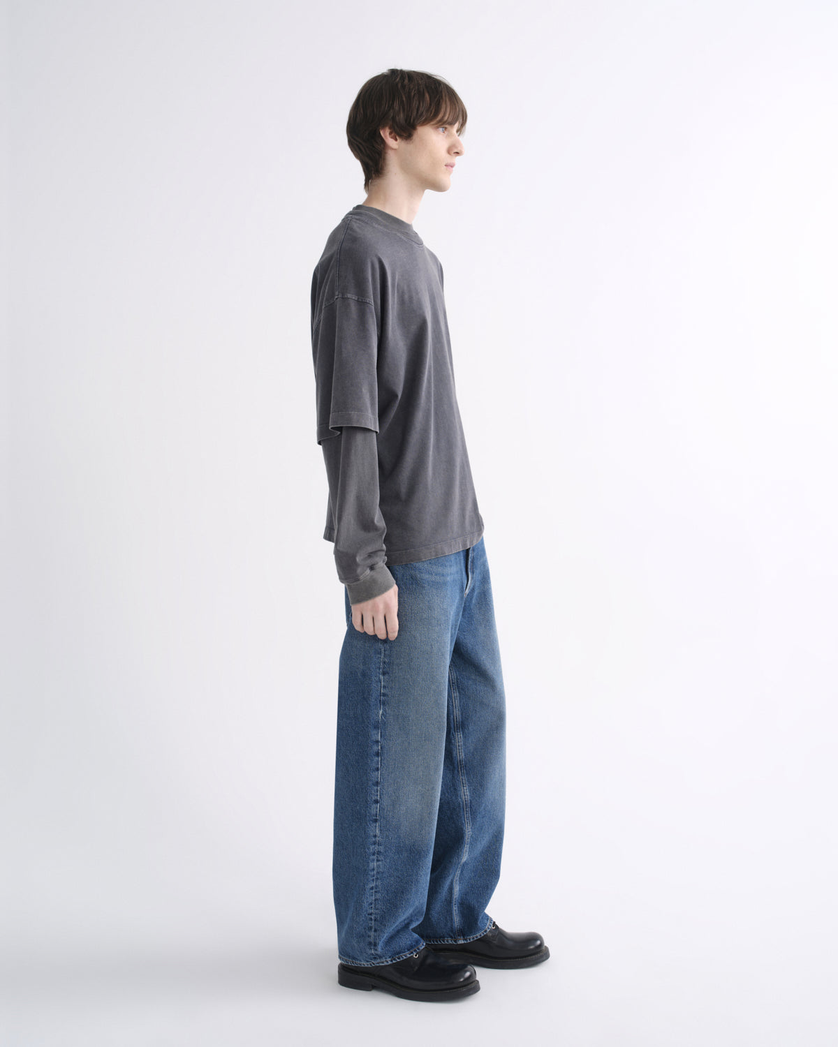 Person wearing a dark grey short sleeve T-shirt layered over a black long sleeve shirt with blue jeans and black shoes, side view, on a grey background
