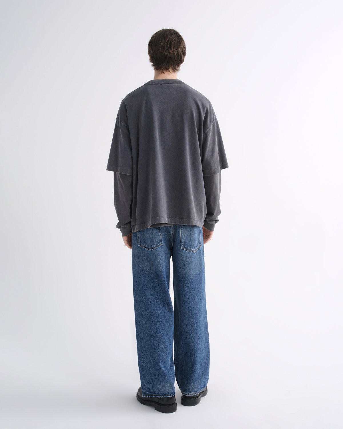 Person wearing a dark grey short sleeve T-shirt layered over a black long sleeve shirt with blue jeans and black shoes, back view, on a grey background