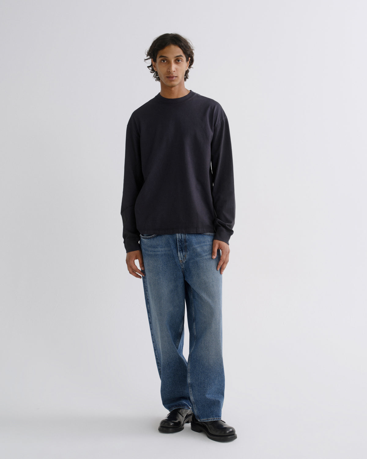 A man stands against a light gray studio background wearing a dark navy long-sleeve crewneck sweatshirt, blue jeans, and black shoes.