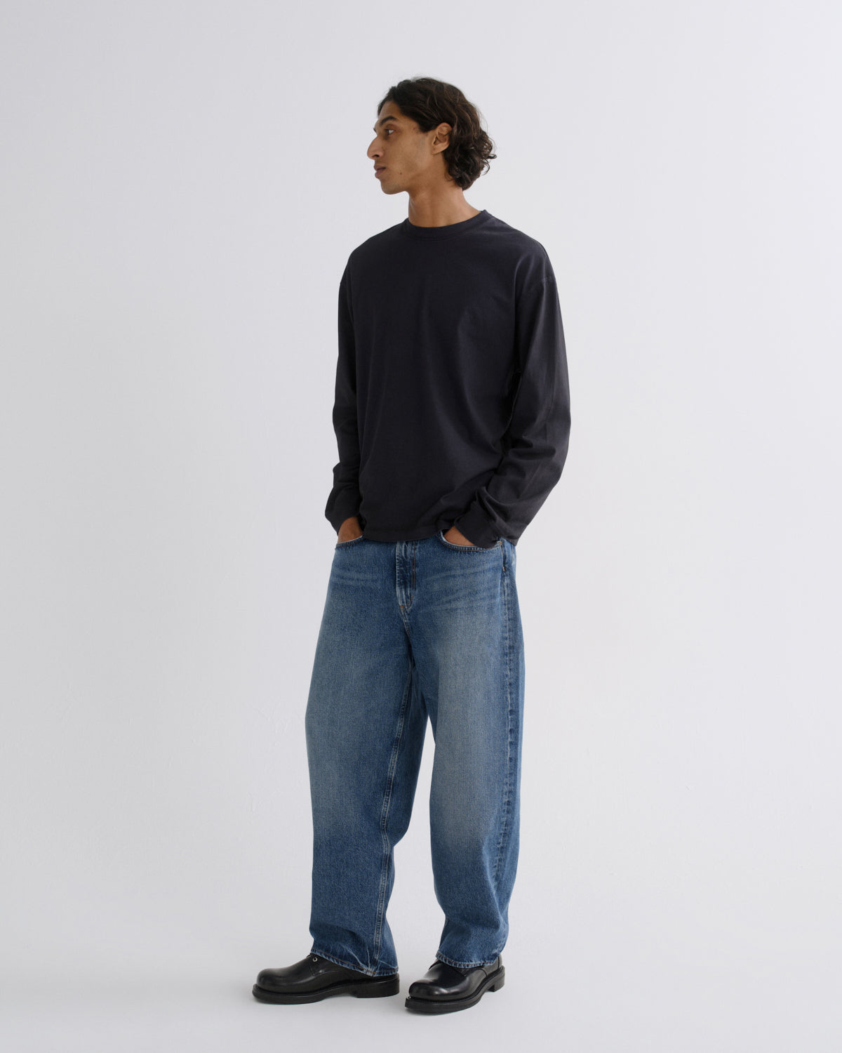 A man stands in profile wearing a dark navy long-sleeve crewneck sweatshirt, loose-fit medium-wash blue jeans, and black leather shoes against a plain light gray studio background.