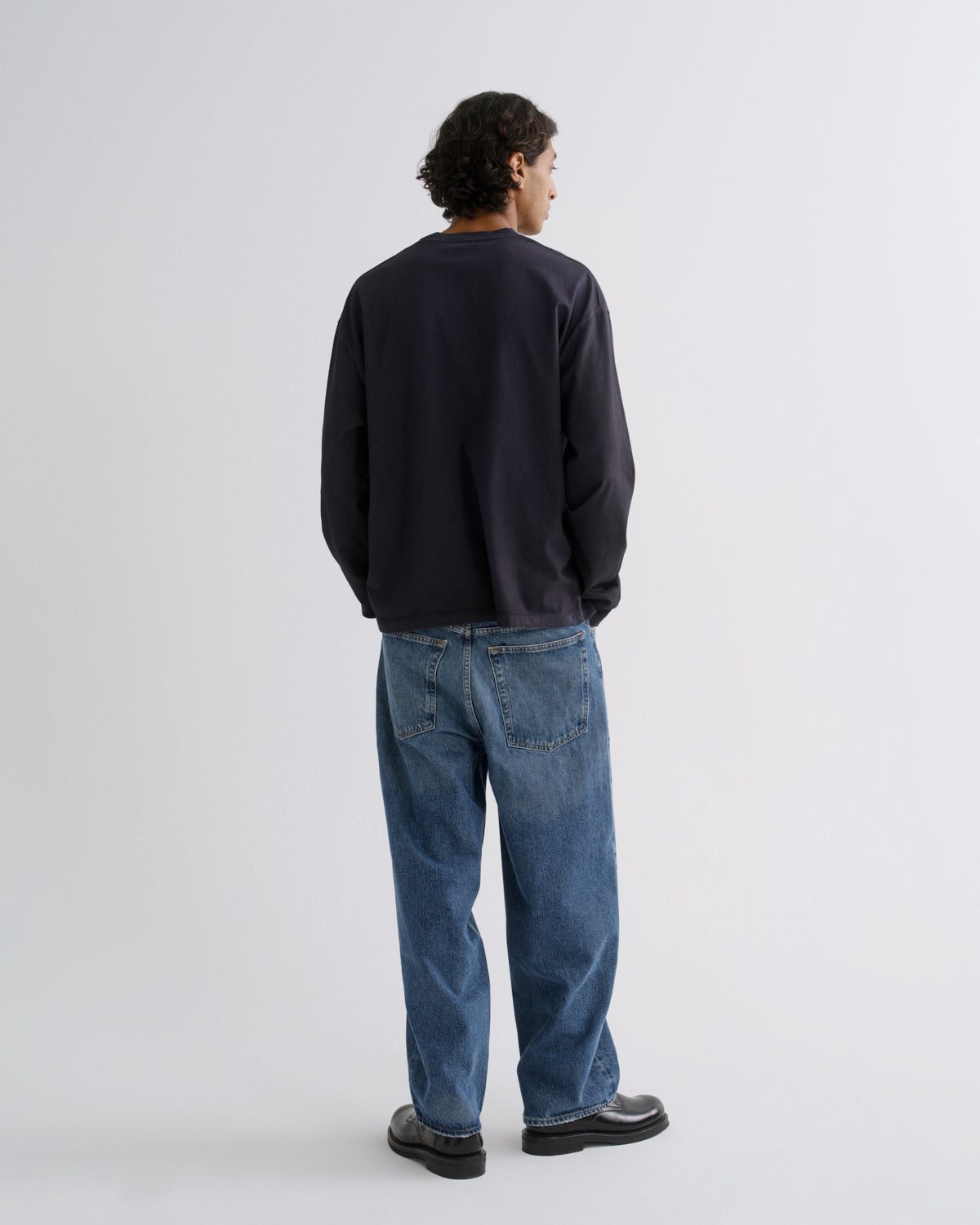 A man viewed from behind wearing a dark navy crewneck sweatshirt, blue jeans, and black shoes, standing against a light gray studio background.