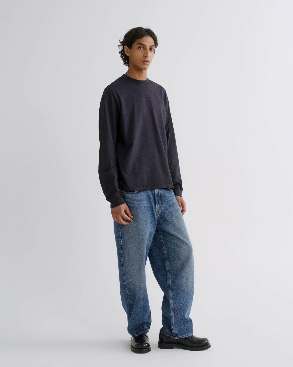 A model stands in a studio wearing a dark charcoal crewneck sweatshirt, loose-fit blue jeans, and black leather shoes against a light gray background.