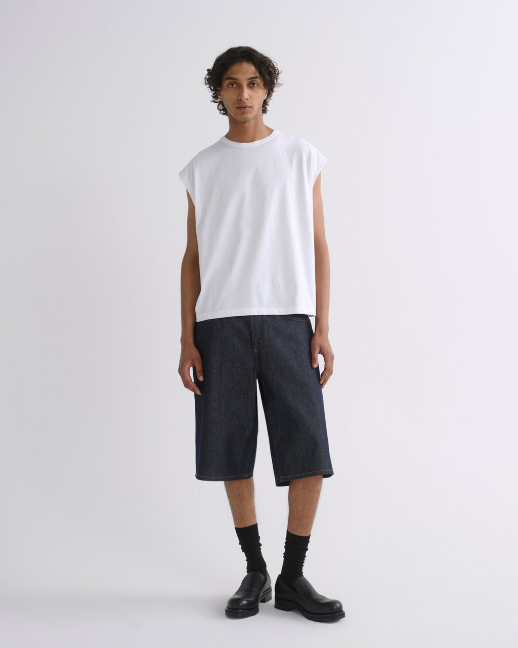 A model stands against a light gray studio background wearing a white sleeveless t-shirt, dark indigo denim shorts, black socks, and black leather boots.