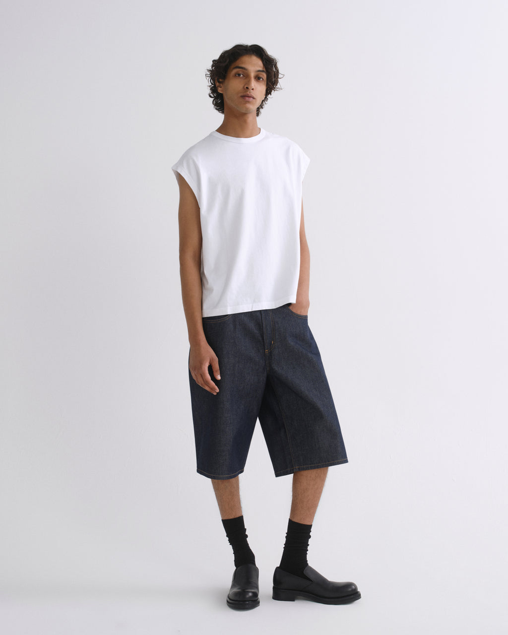 A model stands against a light gray studio background wearing a white sleeveless crewneck t-shirt, dark indigo denim shorts, black socks, and black leather boots.