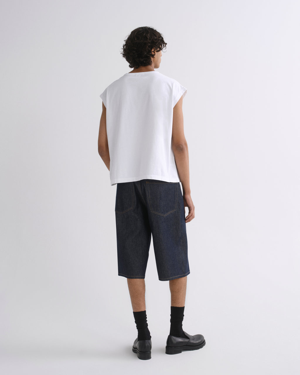 A model shown from the back wearing a white sleeveless t-shirt, dark blue denim shorts, black socks, and black leather shoes, set against a plain light gray studio background.