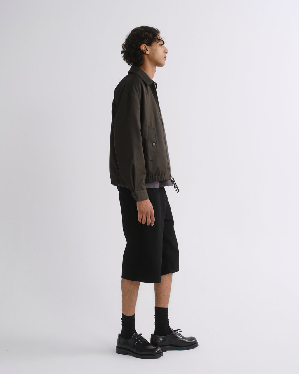 A man in side profile wearing a dark olive-brown utility jacket, black shorts, black socks, and black leather shoes, standing against a light gray studio background.