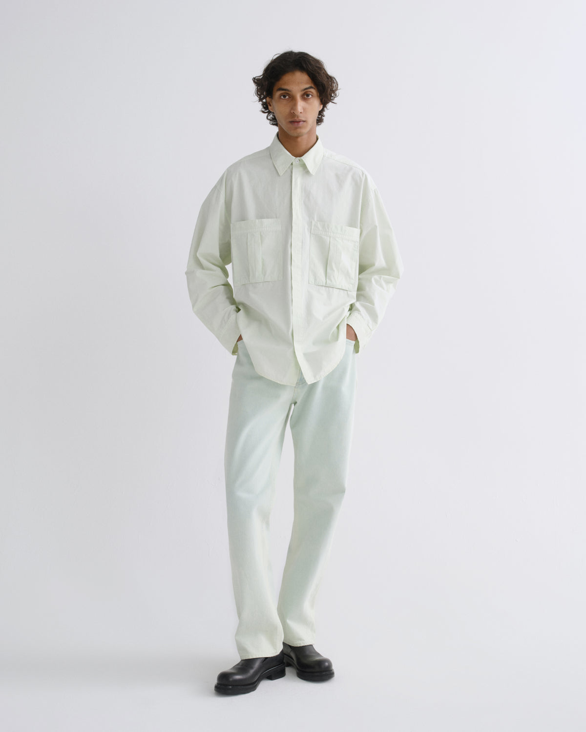A model stands against a light gray studio background wearing a pale mint green long-sleeve button-up shirt with two chest pockets, matching light mint green trousers, and black leather shoes.