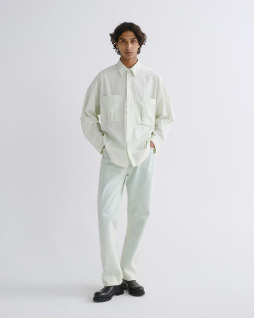 A model stands against a light gray studio background wearing a pale mint green long-sleeve button-up shirt with two chest pockets, matching light mint green trousers, and black leather shoes.