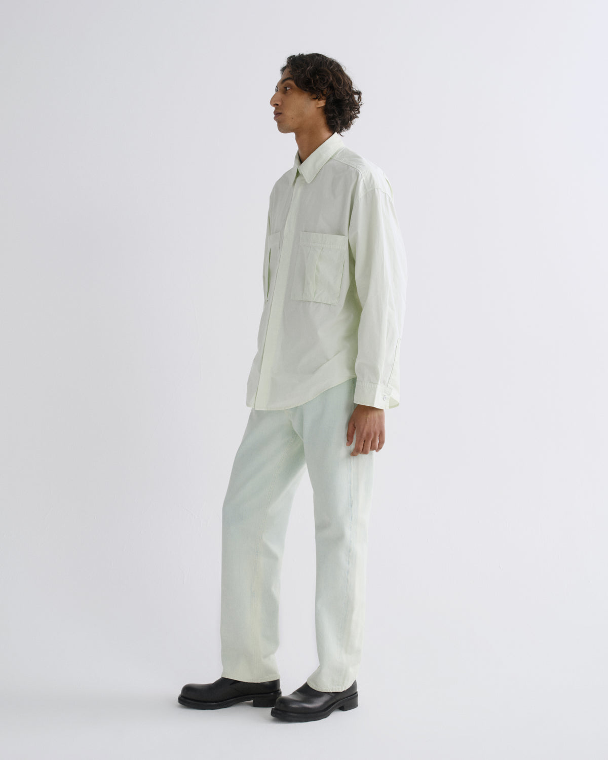 A man stands in profile wearing a light sage green long-sleeve button-up shirt and matching light sage green trousers, paired with black shoes, against a plain light gray studio background.