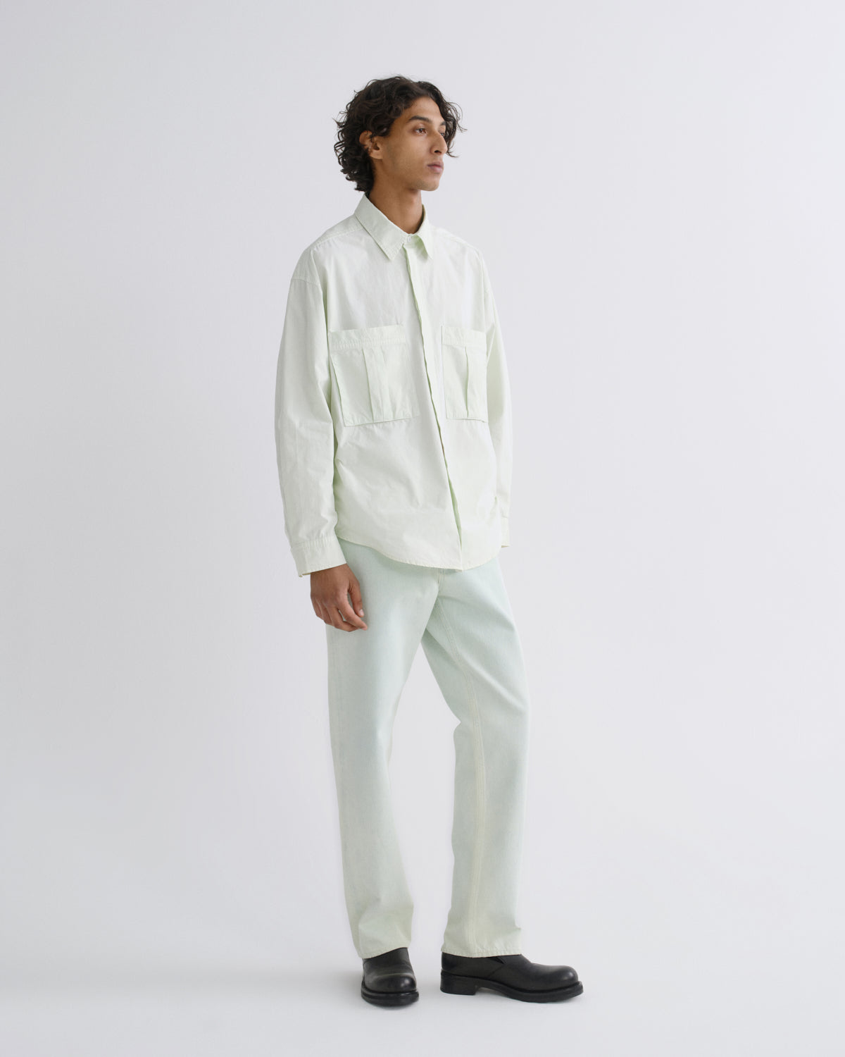A man wearing a light cream long-sleeve button-up shirt with two chest pockets and matching light cream trousers, paired with black shoes, standing against a plain light gray studio background.
