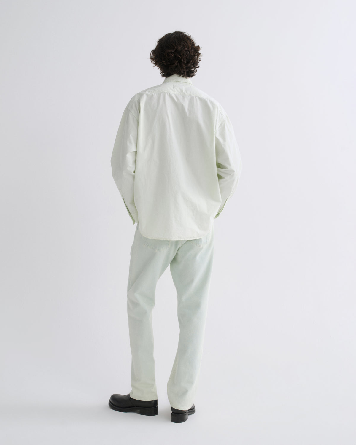 A man viewed from behind wearing a light sage green button-down shirt and matching light sage green trousers, paired with black shoes, against a plain white studio background.