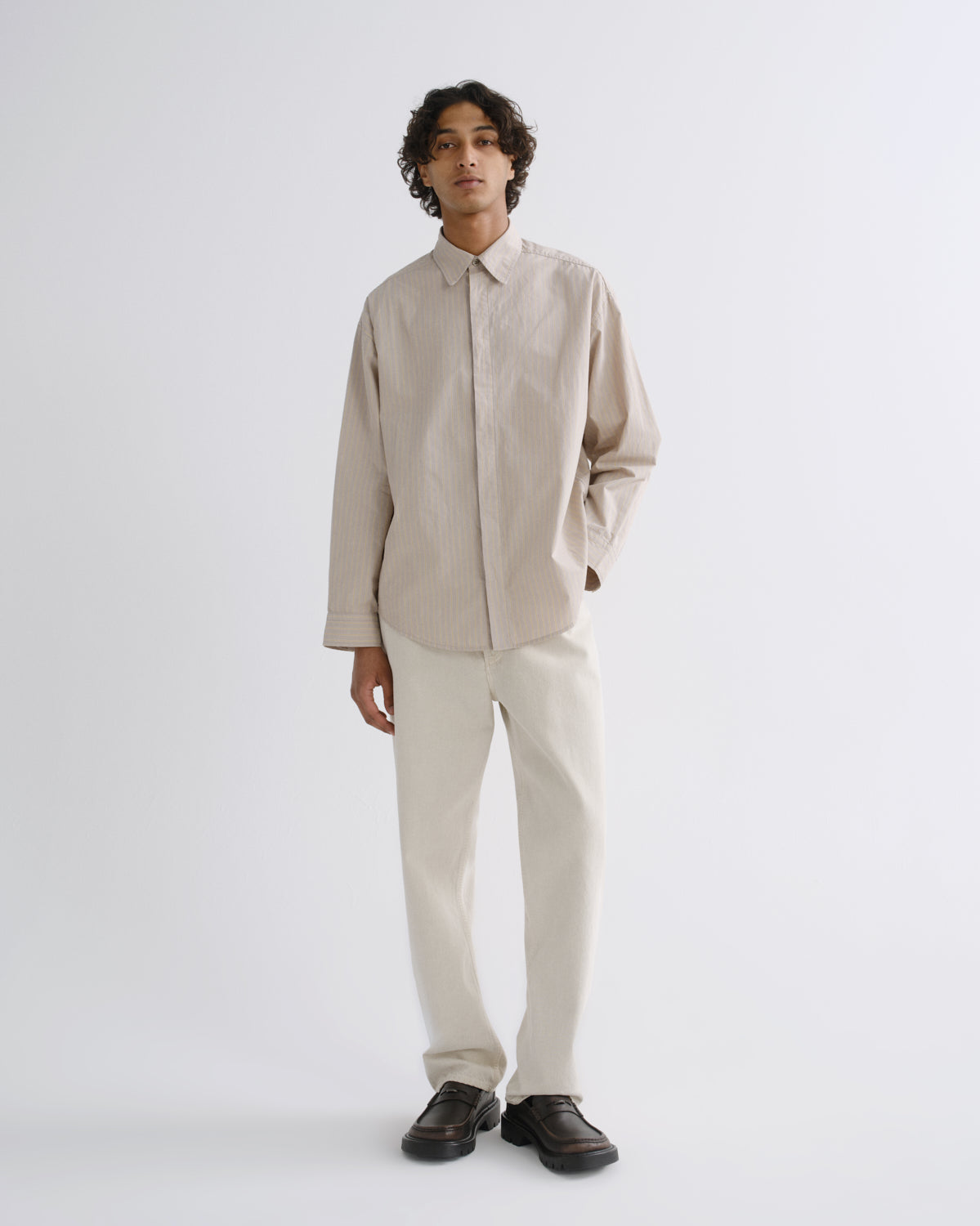 A man modeling a light beige long-sleeve linen button-up shirt and off-white trousers, paired with black loafers, against a plain light gray studio background.