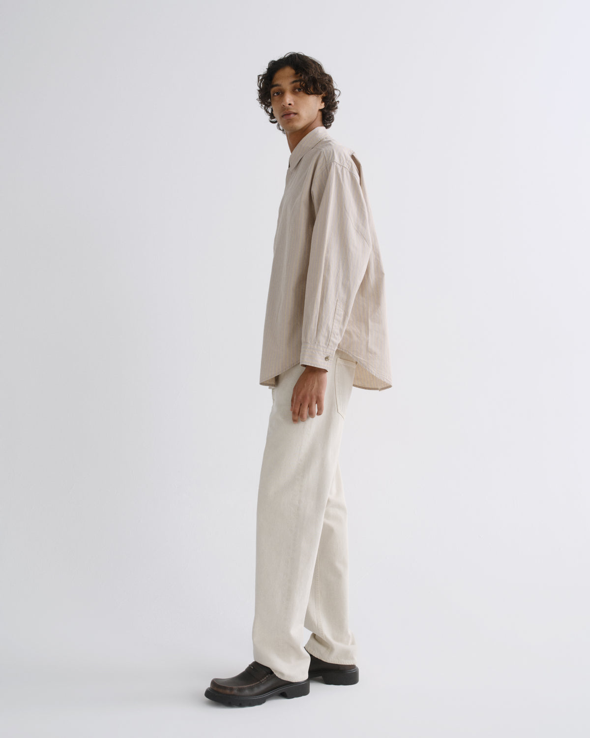A model stands in profile wearing a light beige linen shirt with a unique cutout detail on the back, matching cream linen trousers, and dark brown leather shoes against a plain white studio background.