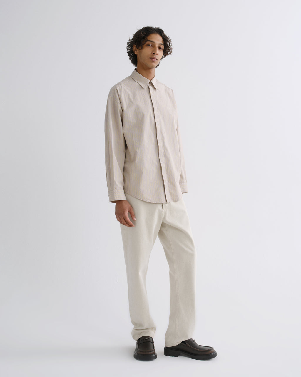 A man modeling a light beige long-sleeve button-up shirt and matching beige trousers, paired with dark brown leather loafers, against a plain light gray studio background.