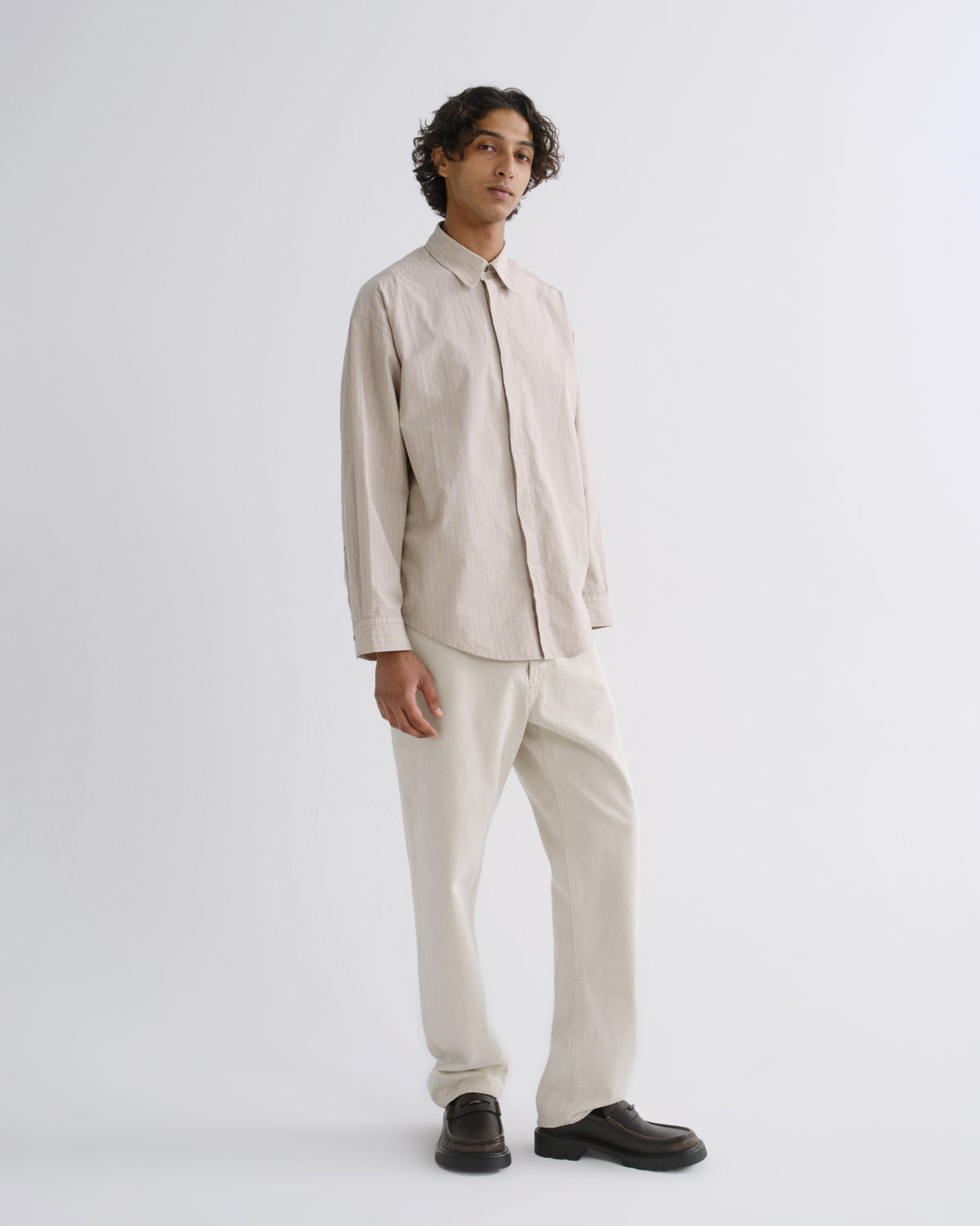 A man modeling a light beige long-sleeve button-up shirt and matching beige trousers, paired with dark brown leather loafers, against a plain light gray studio background.