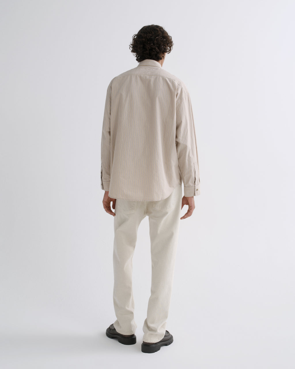 A man viewed from behind wearing a light beige long-sleeve button-up shirt and matching light beige trousers, standing against a plain light gray studio background.
