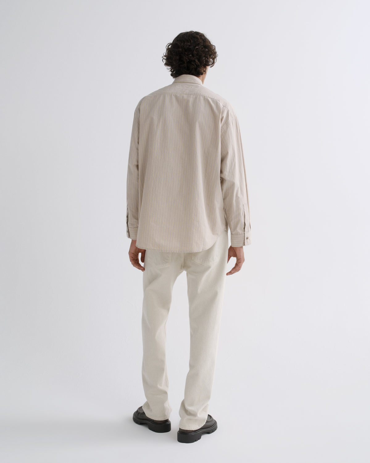 A man viewed from behind wearing a light beige long-sleeve button-up shirt and matching light beige trousers, standing against a plain light gray studio background.
