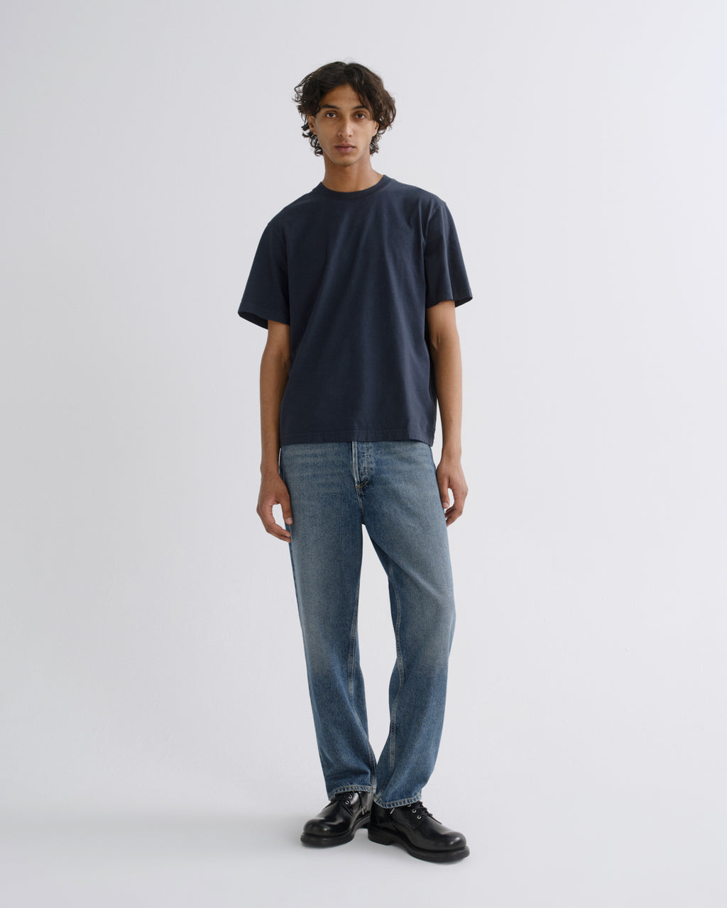 A man stands against a light gray studio background wearing a dark navy short-sleeve crewneck t-shirt, blue jeans, and black leather shoes.