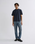 A man stands against a light gray studio background wearing a dark navy short-sleeve crewneck t-shirt, blue jeans, and black leather shoes.