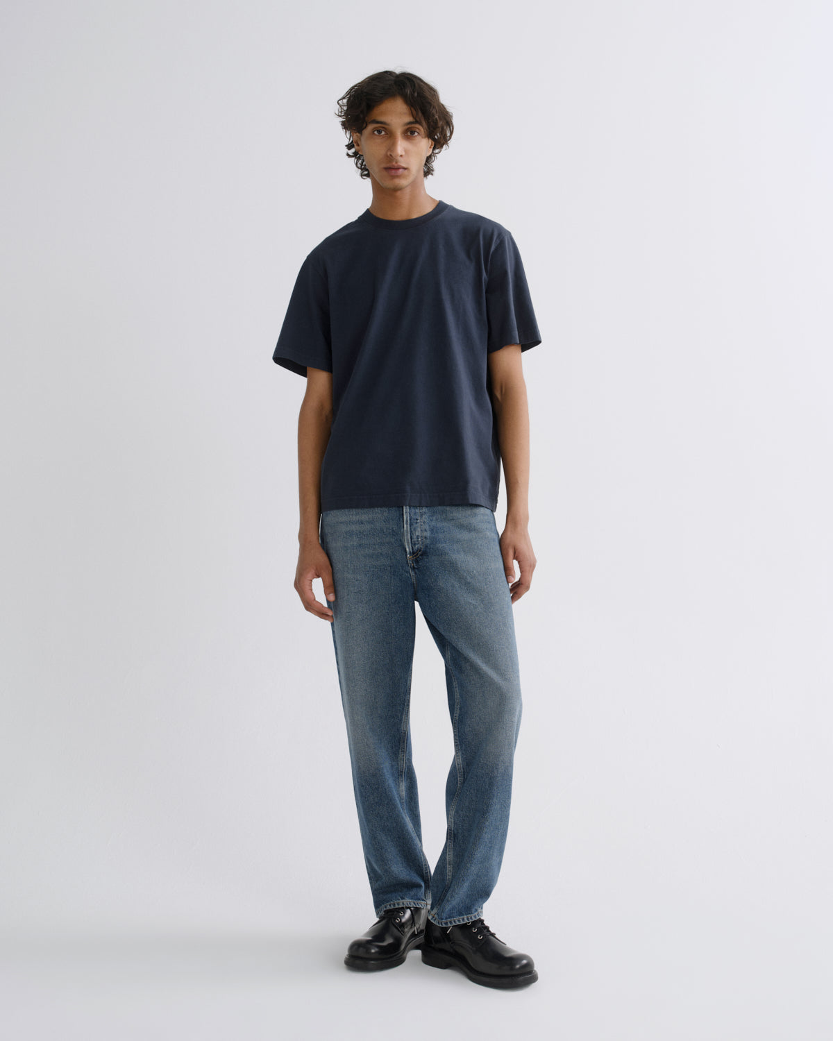A man stands against a light gray studio background wearing a dark navy short-sleeve crewneck t-shirt, blue jeans, and black leather shoes.