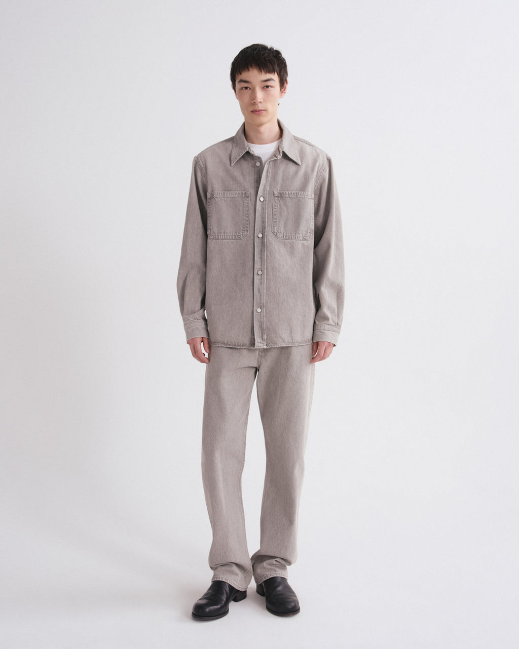 A model stands against a light gray studio background wearing a light gray corduroy button-up shirt jacket with chest pockets, matching light gray corduroy trousers, a white undershirt, and black leather shoes.