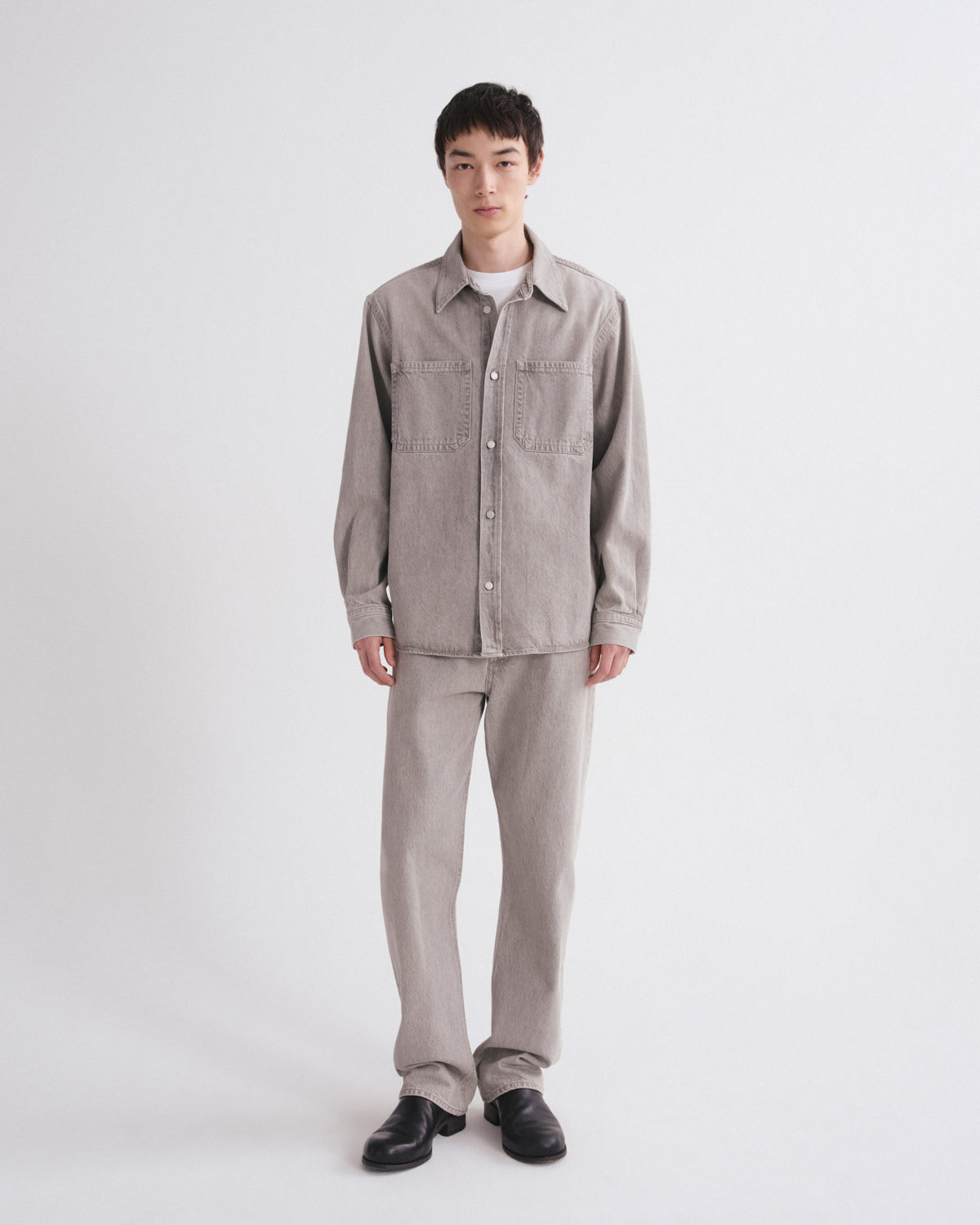 A model stands against a light gray studio background wearing a light gray corduroy button-up shirt jacket with chest pockets, matching light gray corduroy trousers, a white undershirt, and black leather shoes.