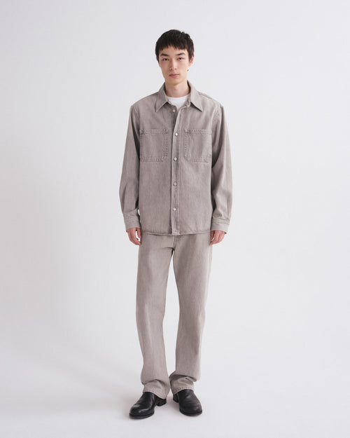 A model stands against a light gray studio background wearing a light gray corduroy button-up shirt jacket with chest pockets, matching light gray corduroy trousers, a white undershirt, and black leather shoes.