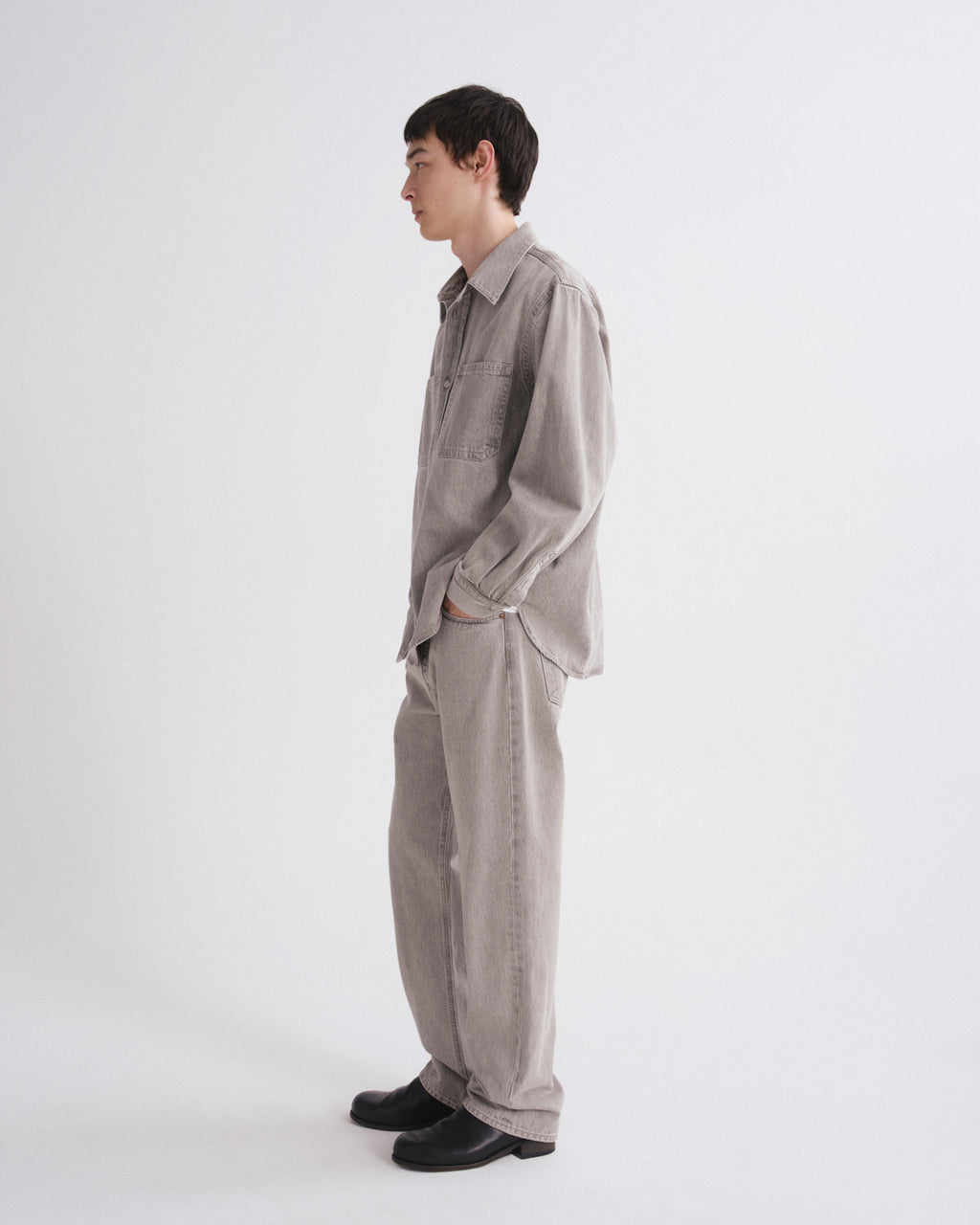 A man in a light gray linen shirt and matching wide-leg trousers, standing in profile against a plain white studio background.