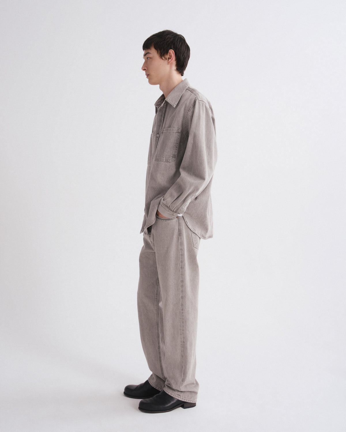 A man in a light gray linen shirt and matching wide-leg trousers, standing in profile against a plain white studio background.