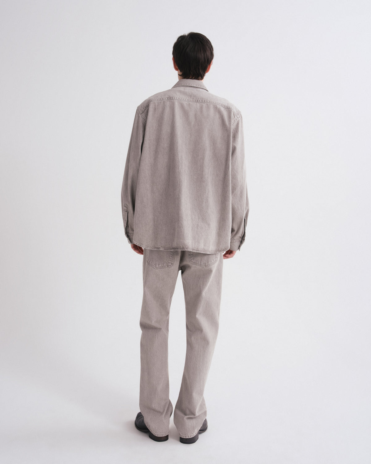 A man viewed from behind wearing a light gray long-sleeve button-up shirt and matching light gray trousers, standing against a plain white studio background.