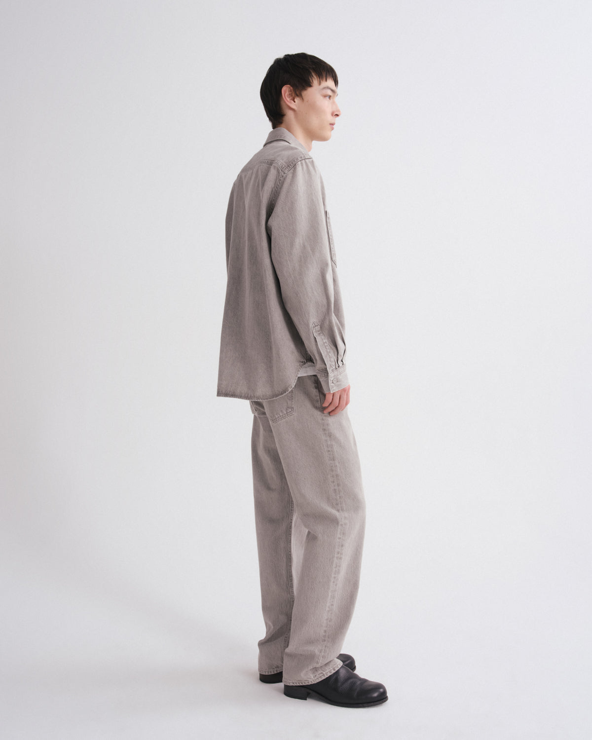 A model shown from a side profile wearing a light taupe-gray corduroy shirt and matching wide-leg trousers, paired with black loafers, against a plain white studio background.