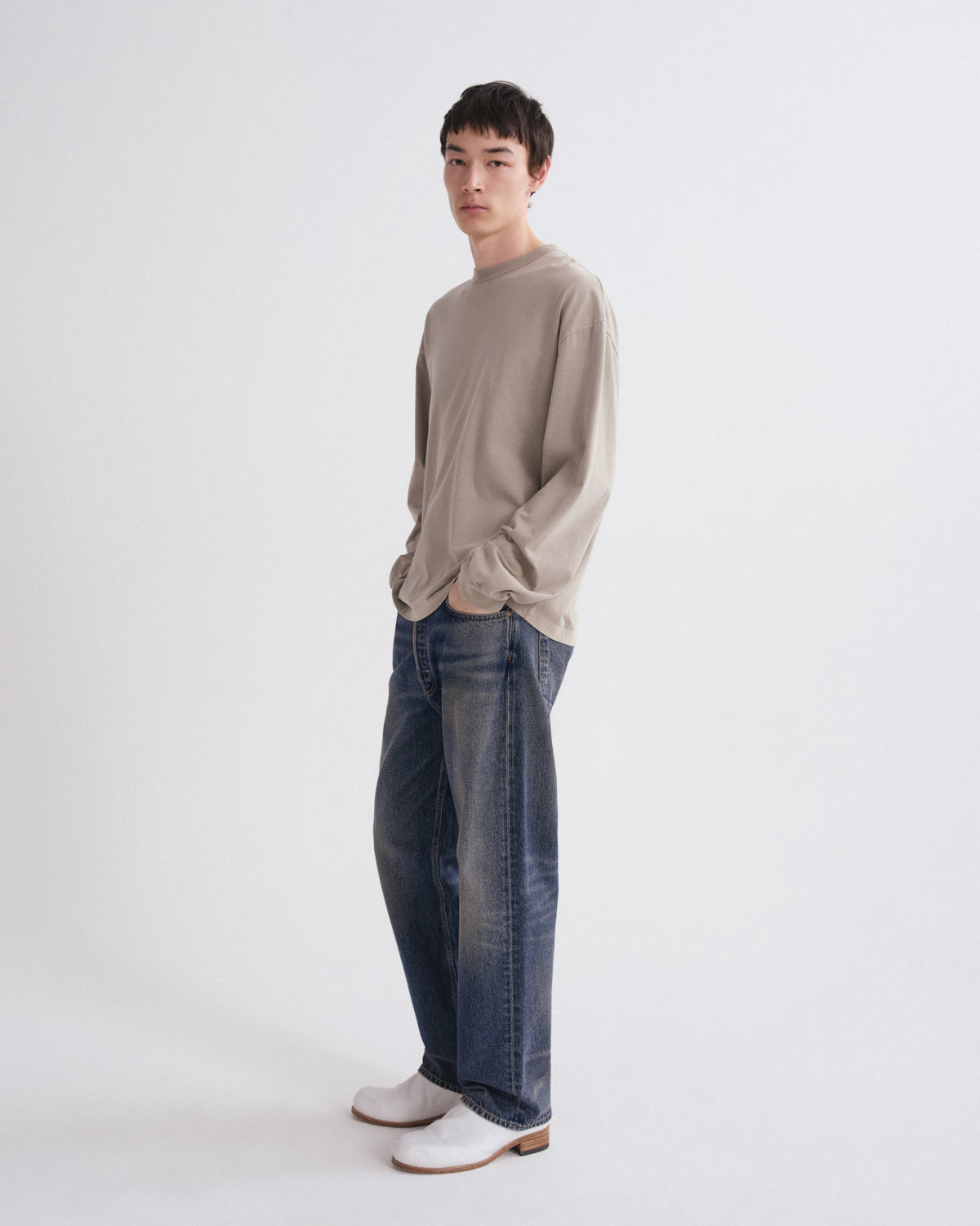 A model stands in profile wearing a light beige long-sleeve knit top, dark blue wide-leg jeans, and white clogs against a plain light gray studio background.