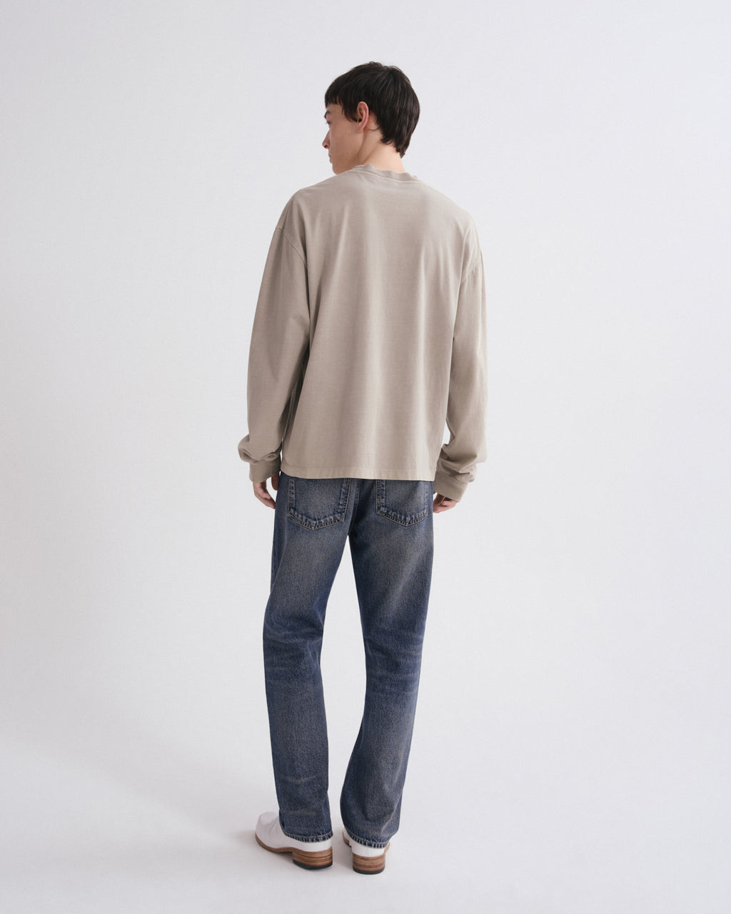 A man is shown from behind wearing a beige long-sleeve crewneck sweatshirt, blue jeans, and white sneakers against a plain white studio background.
