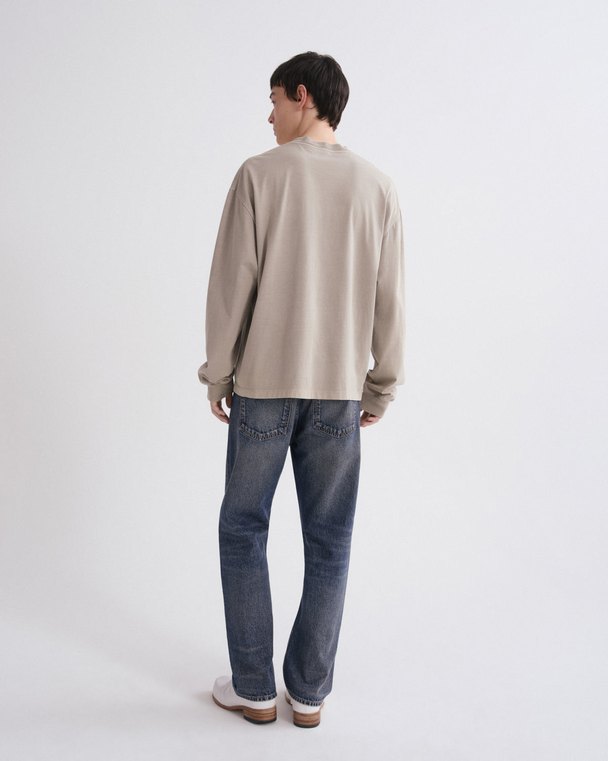 A man is shown from behind wearing a beige long-sleeve crewneck sweatshirt, blue jeans, and white sneakers against a plain white studio background.