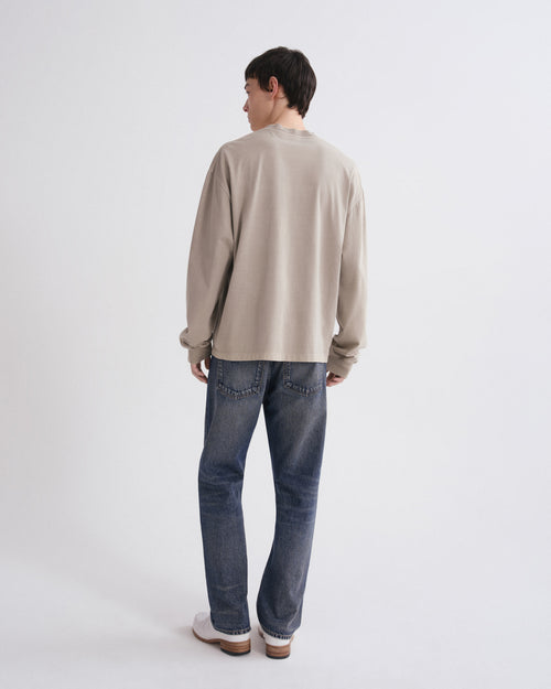 A man is shown from behind wearing a beige long-sleeve crewneck sweatshirt, blue jeans, and white sneakers against a plain white studio background.